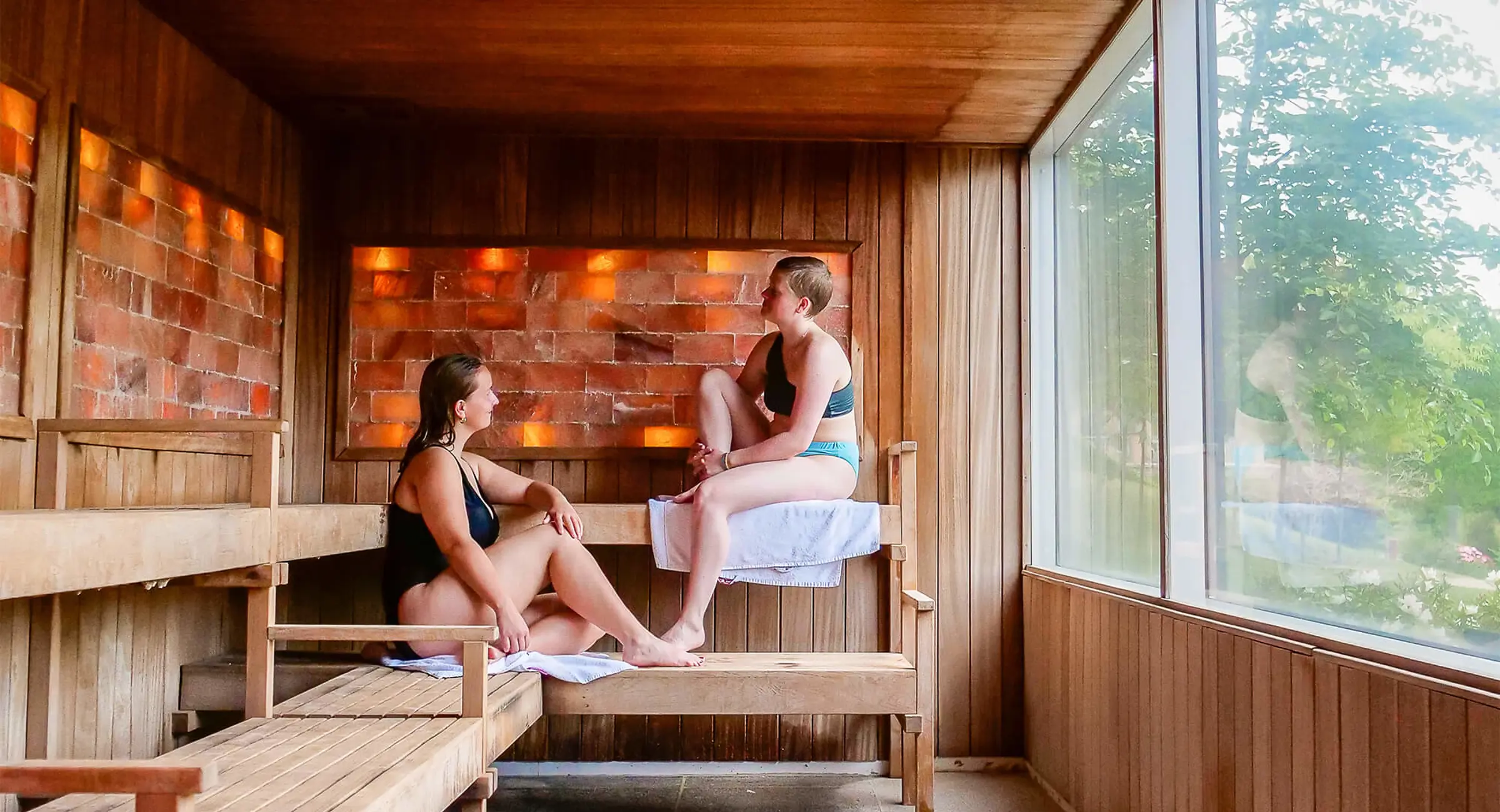 To kvinder i en sauna med himalaya-saltsten på væggen og grønt udenfor vinduet
