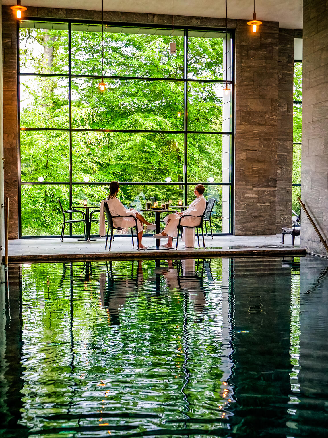 En spa hvor skoven udenfor vinduet spejler sig i vandet. To kvinder sidder ved et bord.