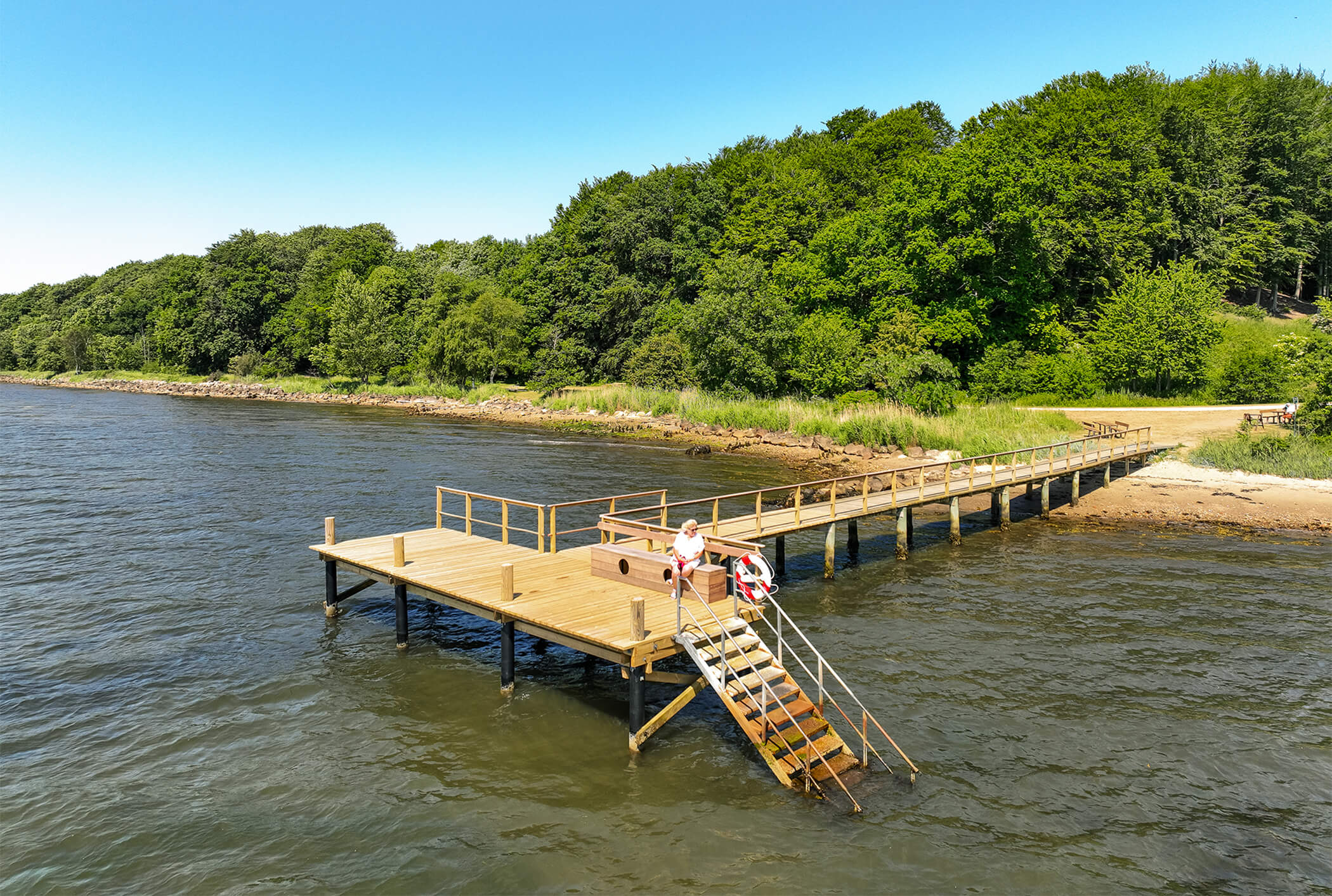 En badebro langs kysten ved Vejle Fjord blå. Blå himmel og grøn skov.