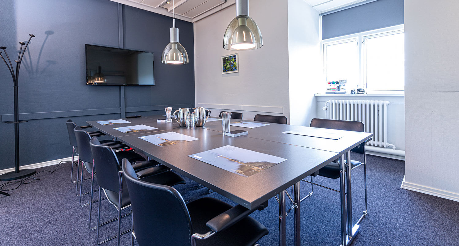 Mødesalon i blågrå farver med bord og stole, stor skærm og vindue