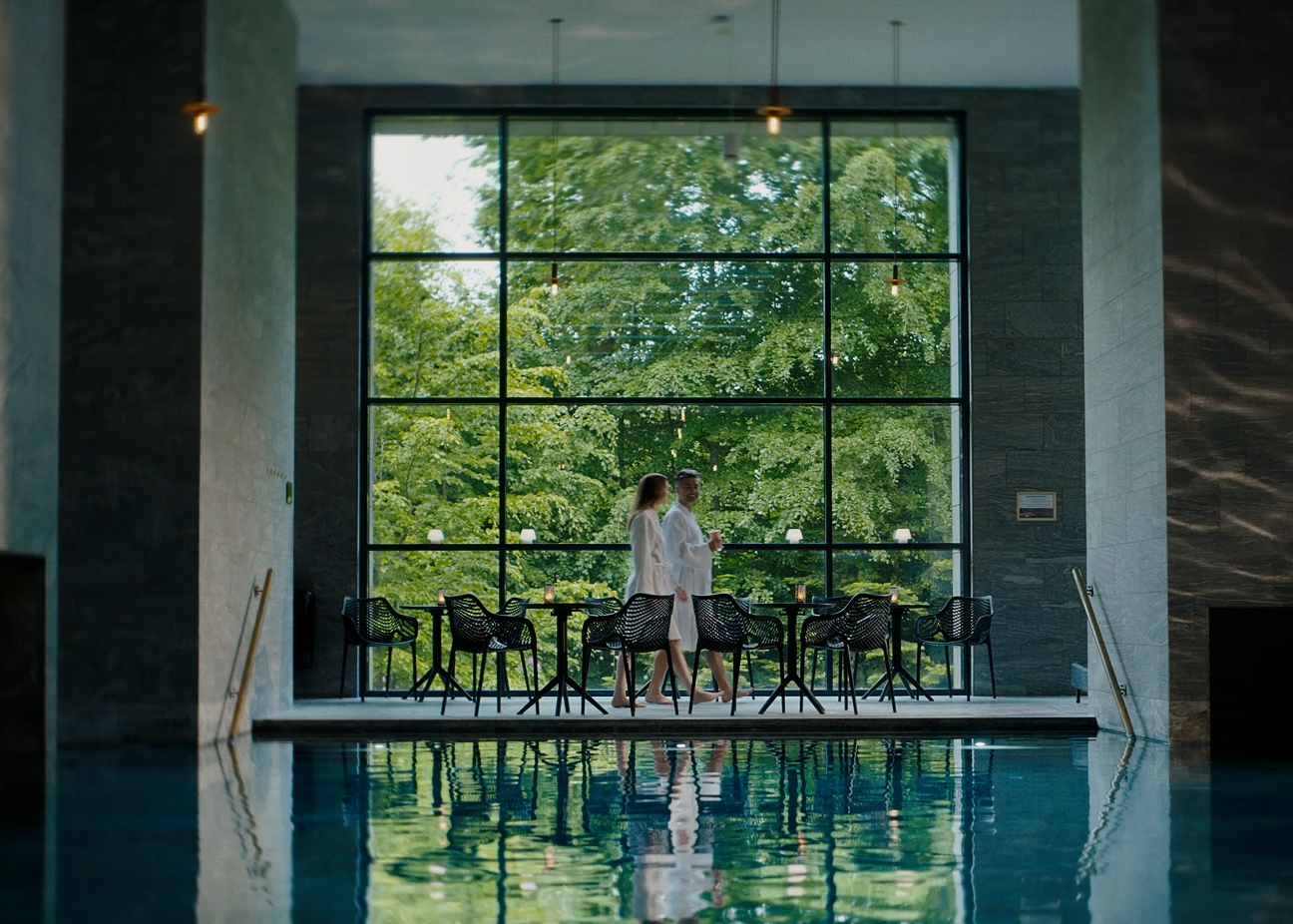 En spa hvor skoven udenfor vinduet spejler sig i vandet. To kvinder sidder ved et bord.