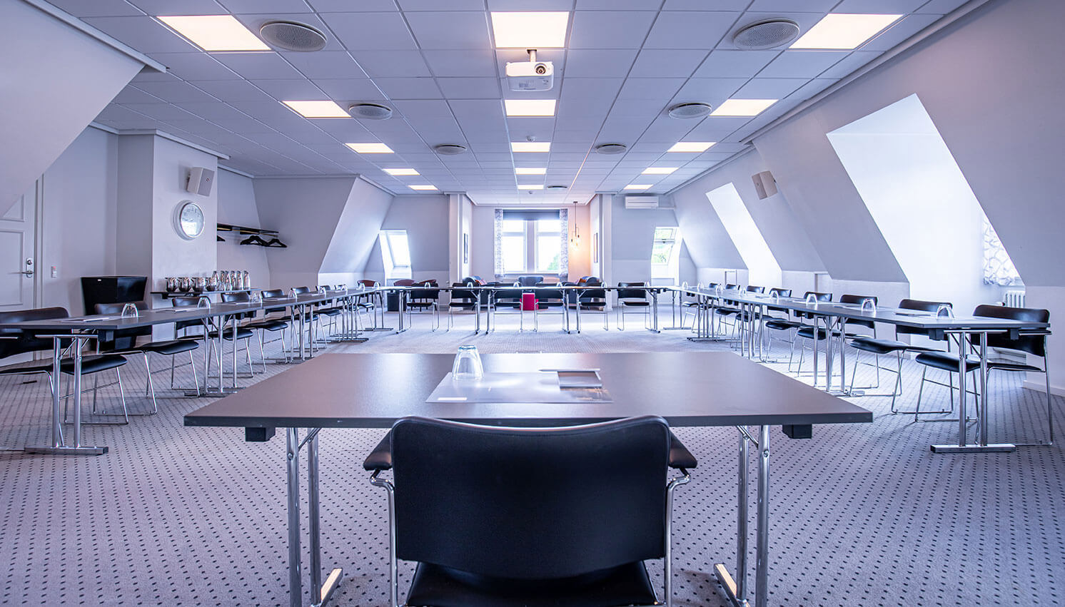 Mødelokale med foredragsholders bord i front og borde opstillet i u-form.