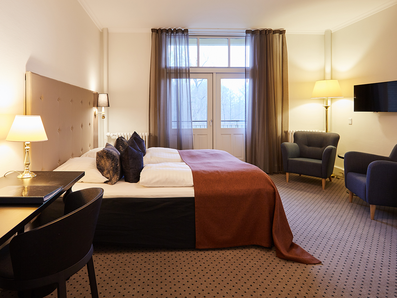 Hotelværelse med dobbeltseng, skrivebord og lænestole og dør til balkon