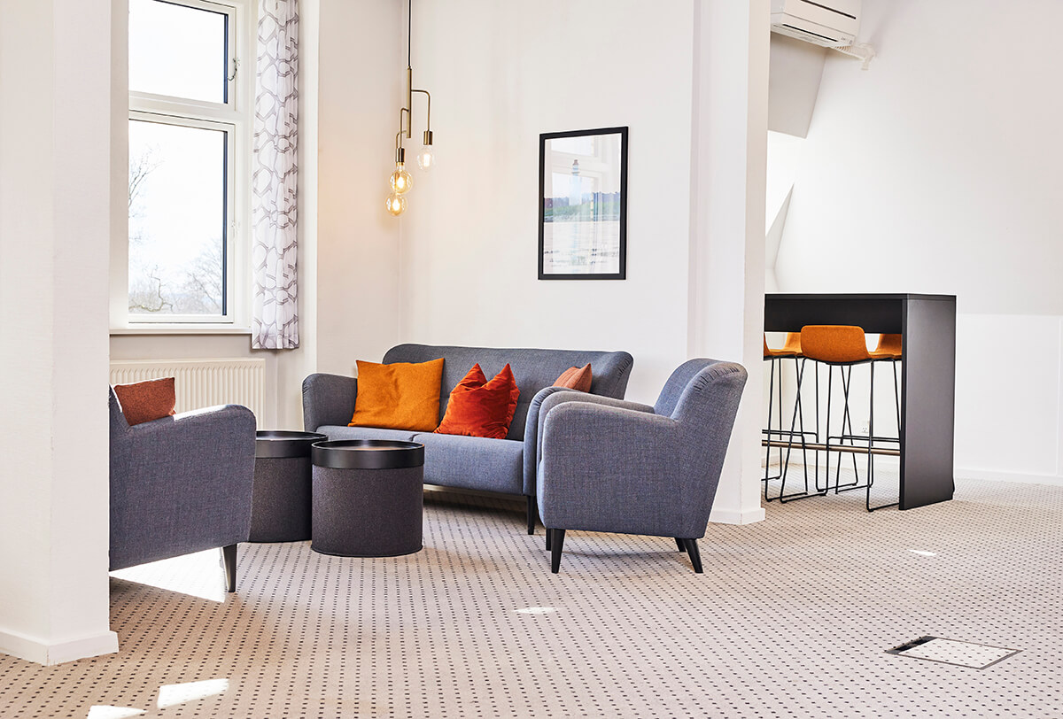 Sofaer og stol med puder ved et vindue, i baggrunden et højt bord med barstole