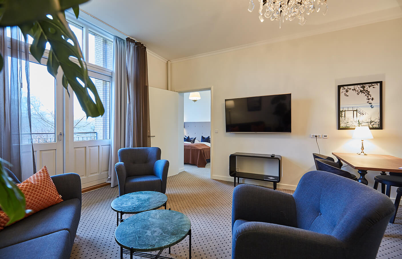 Suite med sofa og lænestole, fjernsyn og balkondøre samt kig til dobbeltseng i andet rum. 