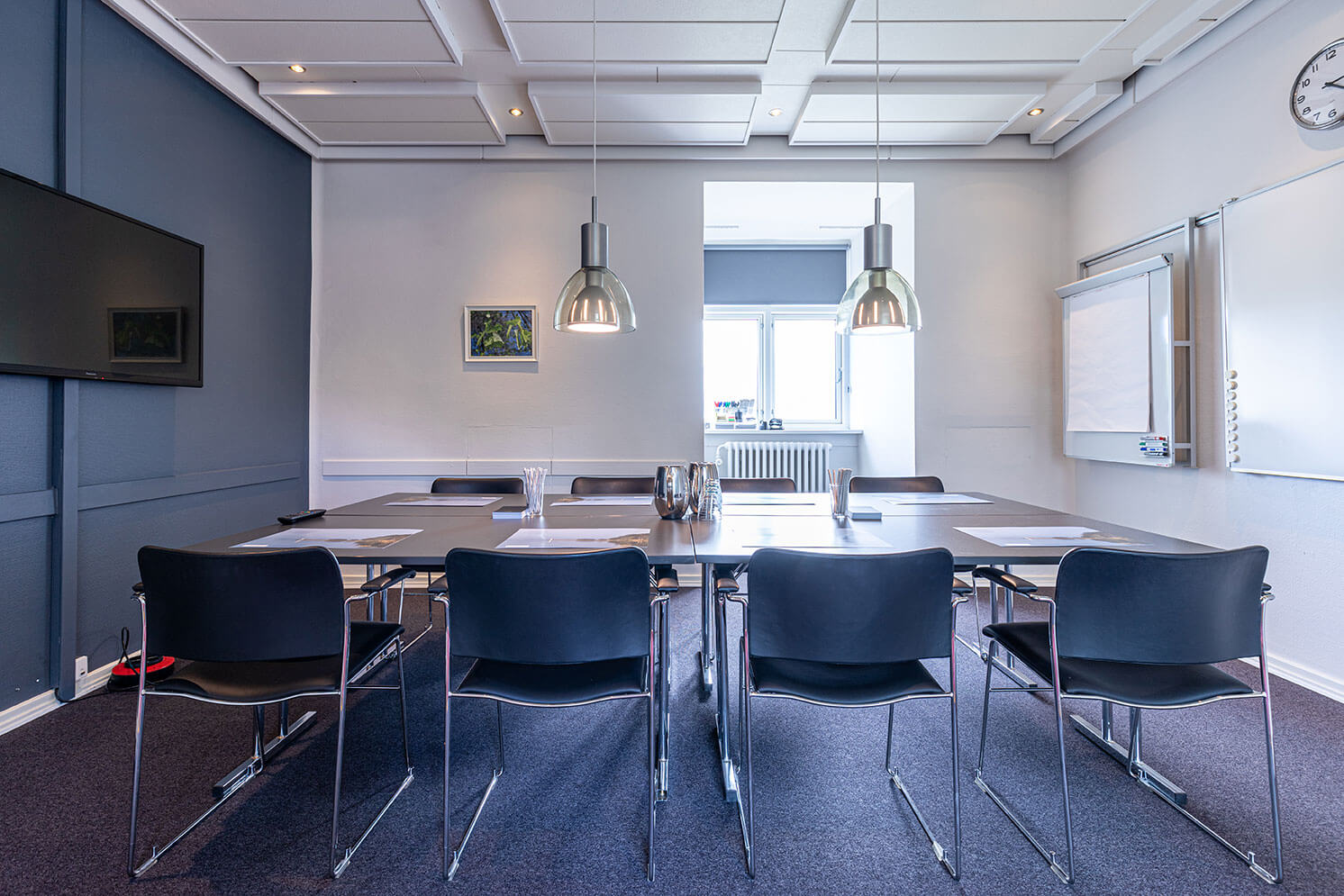 Mødesalon i blågrå farver med bord og stole, stor skærm og vindue